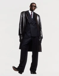 Black model wearing dark suit, leather coat, tie and glasses against a seamless white background for Behind the Blinds.