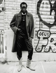 Model wearing a structured coat and trousers posing against a brick wall with graffiti for Behind the Blinds editorial