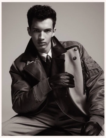 A young male model wearing a paneled bomber jacket, white shirt, tie, and leather gloves against a neutral studio background.