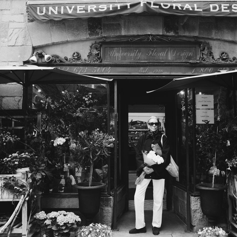 A woman stands in front of University Floral Design with a bouquet of flowers, wearing sunglasses, a jacket, and white pants.
