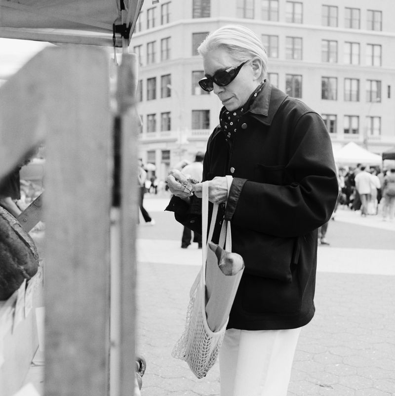 Monochrome medium shot of an adult woman with sunglasses at an outdoor market holding a mesh bag and produce near vendor stands.