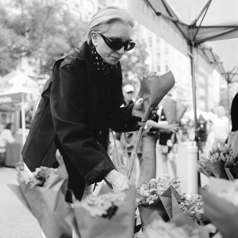 Older woman in sunglasses buying flowers at an outdoor market, in a black-and-white photo with buildings visible in the background.