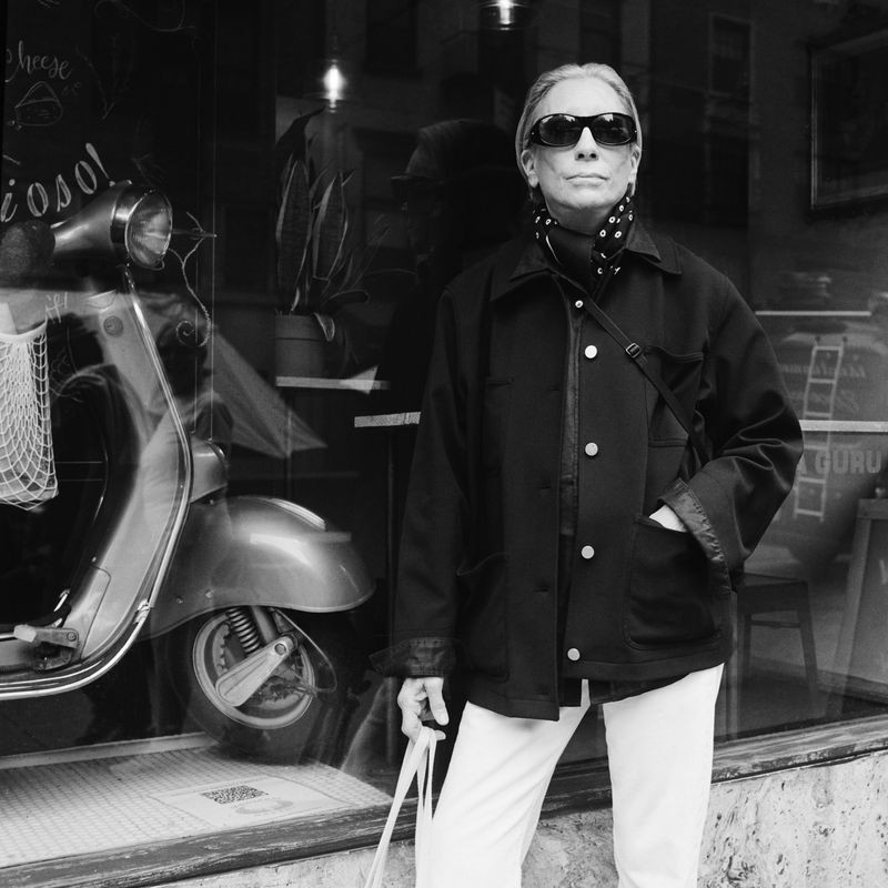 Black and white shot of a woman in sunglasses and a jacket, standing on a street next to a vintage scooter, urban setting.