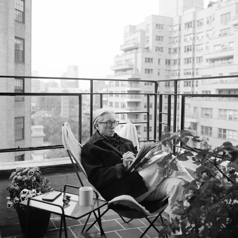 Monochrome photo of an adult male with fair hair, wearing glasses and a coat, sitting in a chair on a balcony reading a newspaper.