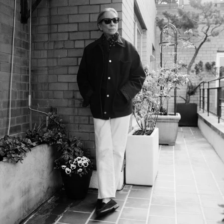 Black and white outdoor shot of a stylish older woman in sunglasses, coat, and white pants, standing on a balcony with potted plants.