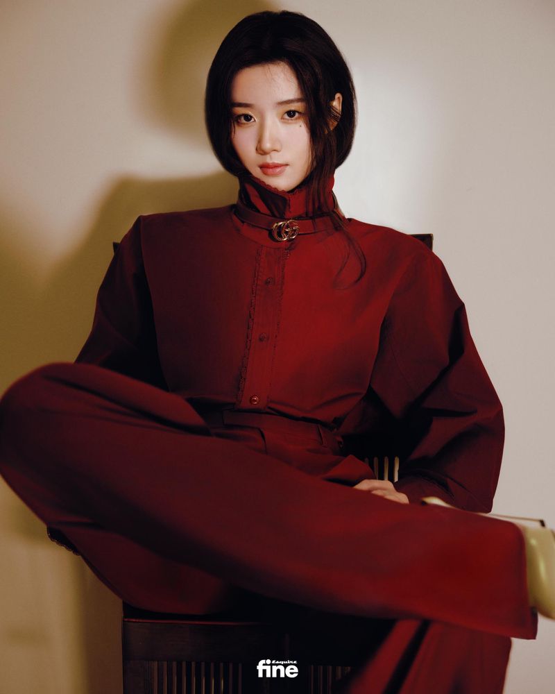 Portrait of a young Asian woman with black hair in a red pantsuit sitting on a dark wood chair in front of a neutral background.