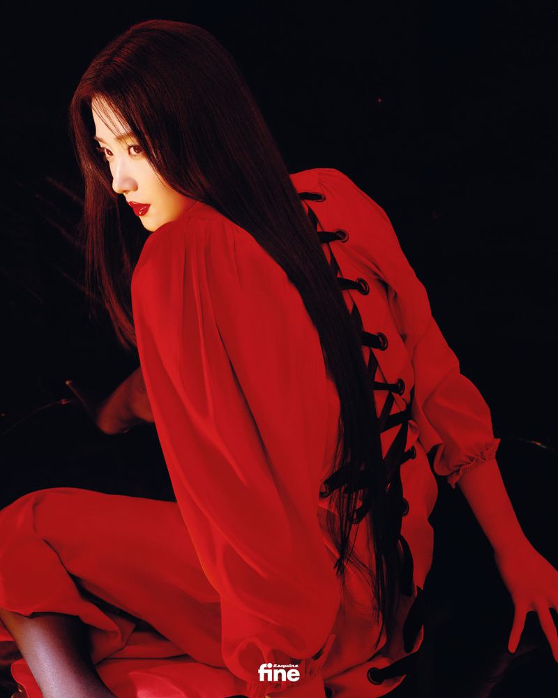 Woman in red ensemble sitting, long hair flowing, against a dark backdrop. Fashion editorial, studio lighting, dramatic red hue.