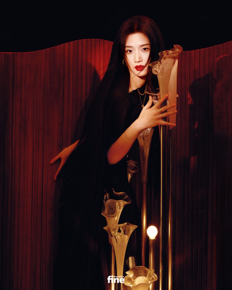 Portrait of a woman with long black hair, red lipstick, and a black dress, standing in front of a red pleated backdrop.