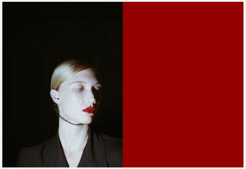 Woman with red lipstick and a net veil poses in front of a split black and red background in a beauty or fashion shot.