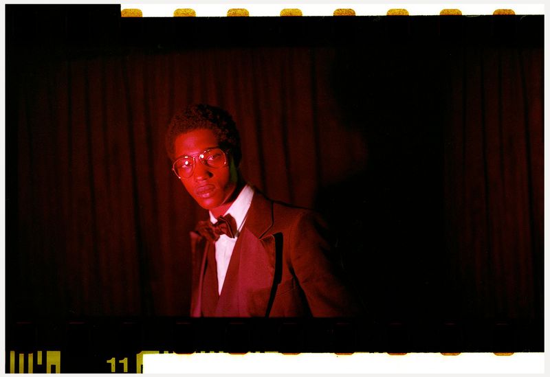 Portrait of an African-American man in a tuxedo with a bow tie and glasses, looking at the camera against a dark red curtain backdrop.