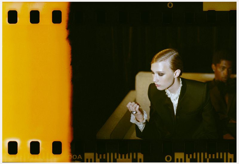 Portrait of a model in a black blazer with lace collar, an orange film border on the left, and a blurry man in the background.