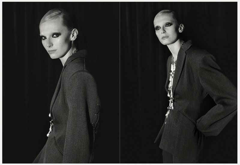Monochrome portrait of a stylish woman in a blazer with bold eye makeup, standing against a dark backdrop in a studio setting.