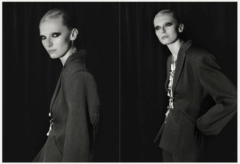 Monochrome portrait of a stylish woman in a blazer with bold eye makeup, standing against a dark backdrop in a studio setting.