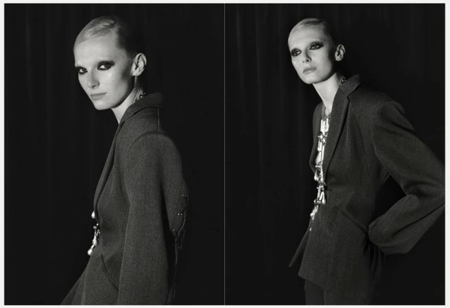 Monochrome portrait of a stylish woman in a blazer with bold eye makeup, standing against a dark backdrop in a studio setting.