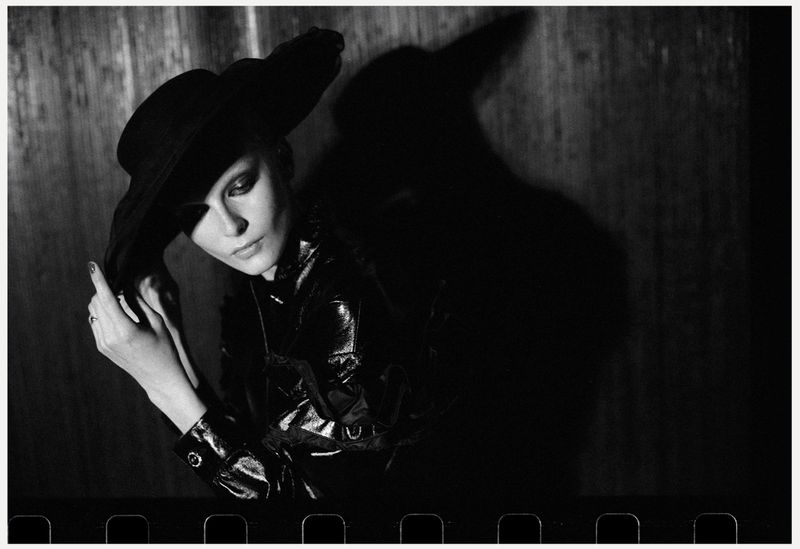 Black and white portrait of a woman in a wide-brimmed hat and shiny jacket in a high-contrast, indoor setting with prominent shadow.