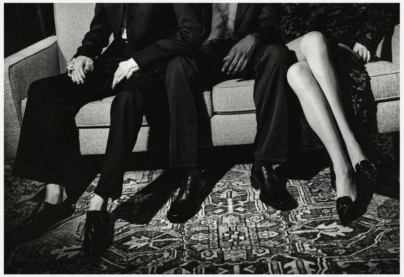 Three people pose while sitting on a couch; two men in dark suits and a woman's legs wearing dark heels and patterned dress.