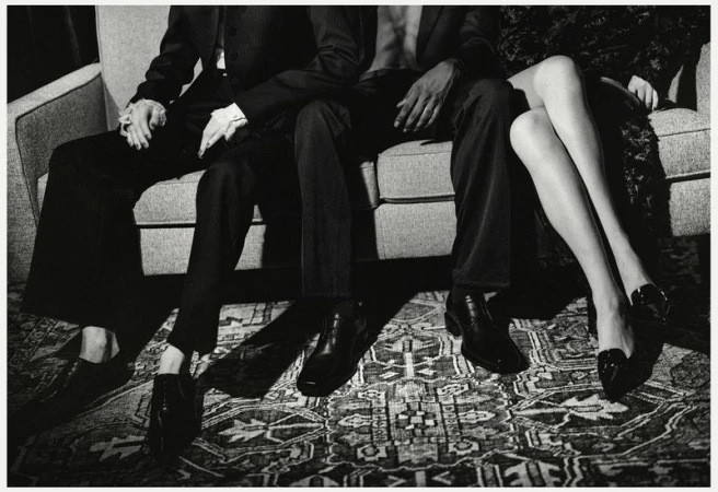 Three people pose while sitting on a couch; two men in dark suits and a woman's legs wearing dark heels and patterned dress.