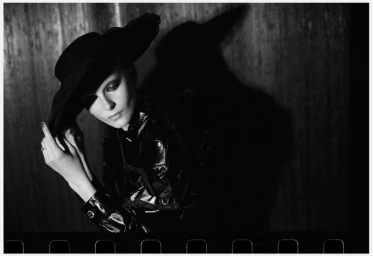 Model wearing a wide-brimmed black hat and dark jacket posing against a textured wall with a dramatic shadow