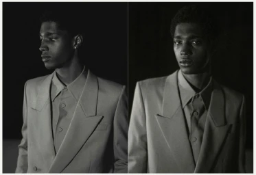 Black and white editorial portrait of a young Black man wearing a buttoned jacket and collared shirt in low key lighting.