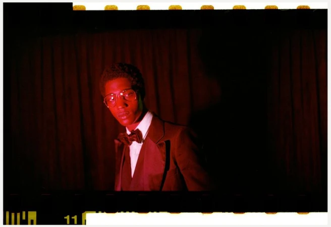 A man wearing glasses and a formal suit jacket with a bowtie against a dark red curtain background in an editorial style portrait.