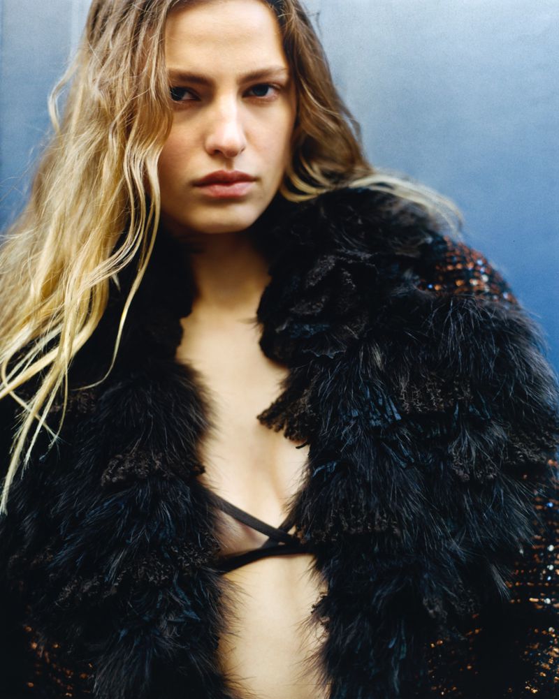 Blonde model with dark feather coat and bra posing against blue backdrop in studio lighting, fashion editorial photography.