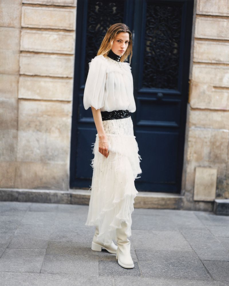 A full shot of a woman in a tiered white dress with long sleeves and white boots, posing against a building with a dark blue door.