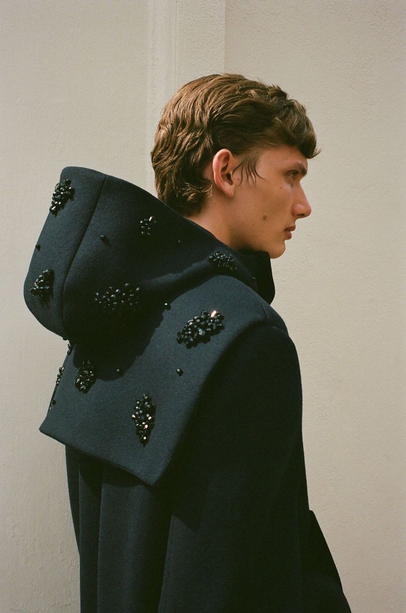 Fashion portrait of a young adult male with dark blond hair, wearing a dramatic black hooded coat with beaded embellishments indoors.