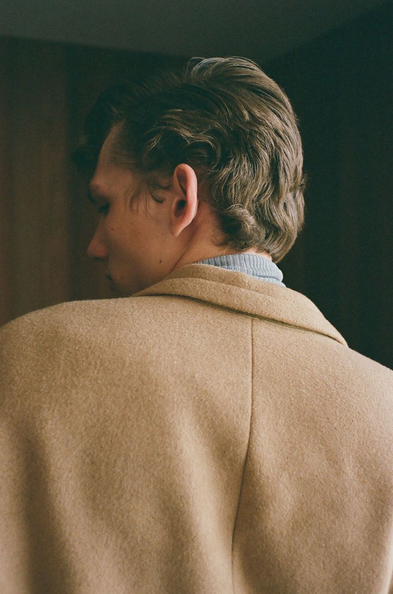 Back view of an adult man with dark hair wearing a tan wool coat and blue sweater indoors. Fashion or lifestyle photography.
