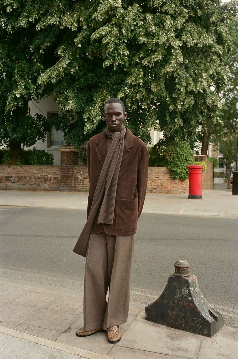 A dark-skinned man in a brown coat stands on a city sidewalk with a tree and a red mailbox in the background on a sunny day.