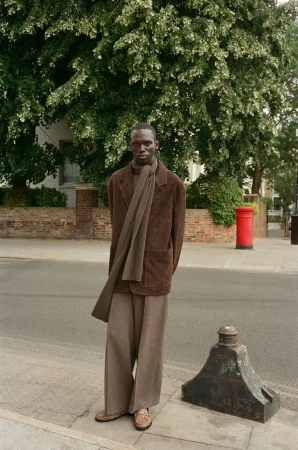 A dark-skinned man in a brown coat stands on a city sidewalk with a tree and a red mailbox in the background on a sunny day.