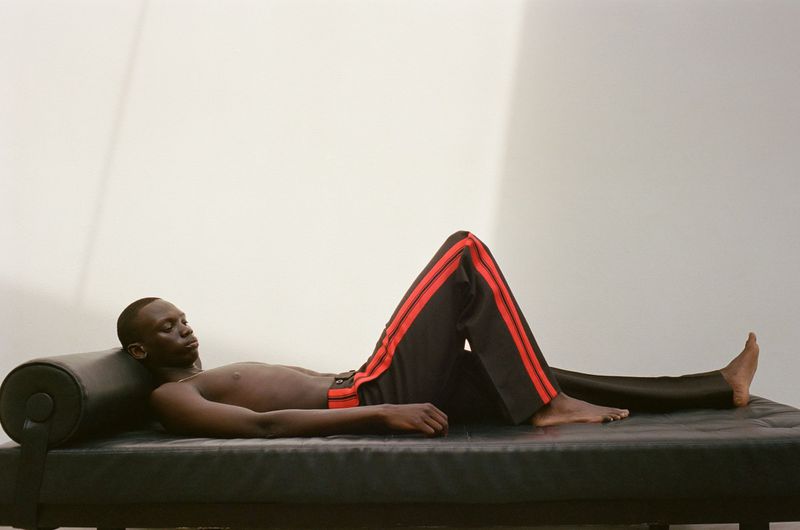 Adult male lying on a black platform wearing black pants with red stripes against a white wall in a photo studio.