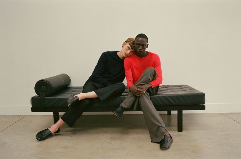Two men pose casually on a leather bench in a studio. One rests his head on the other's shoulder. Minimalist aesthetic with soft, diffused lighting.