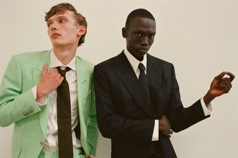 Two adult men pose indoors wearing elegant suits and ties. One man wears a light green suit, while the other wears a black suit.