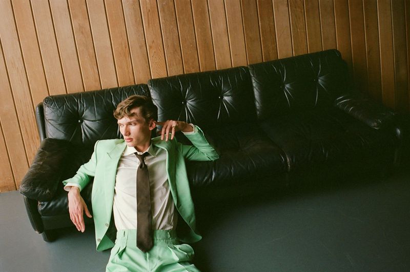 Stylish adult male in a light green suit sits on the floor next to a black leather couch against a wooden wall in a studio setting.