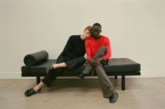 Two male models seated together on a black leather daybed against a plain light wall for The Greatest Magazine editorial.