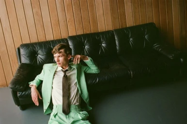 A young male model in a mint green suit and brown tie reclining against a black leather couch for The Greatest Magazine.