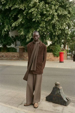 Black man standing on a city street wearing a brown blazer, matching scarf, and trousers, photographed by Clement Pascal.