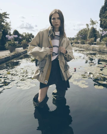 A young woman stands in a body of water, wearing a light-colored jacket, dark skirt, and tall rubber boots, surrounded by greenery.