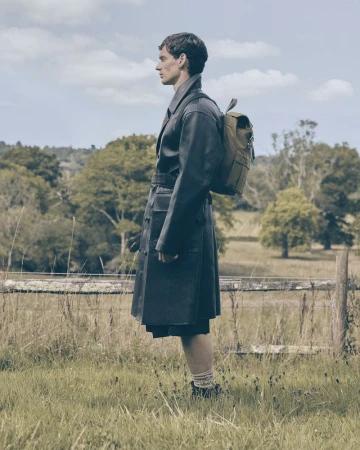 Man in a black coat with a backpack standing in a grassy field under a cloudy sky, looking away, fashion photography, outdoor editorial.