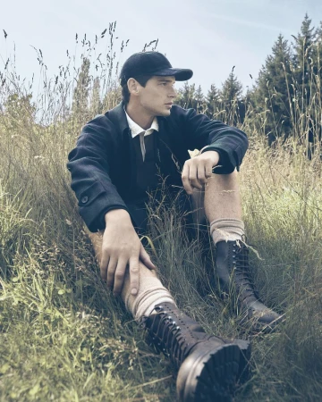 A young adult man sits in a grassy field wearing black lace-up boots, a baseball cap, and knee-high socks against a woodland backdrop.