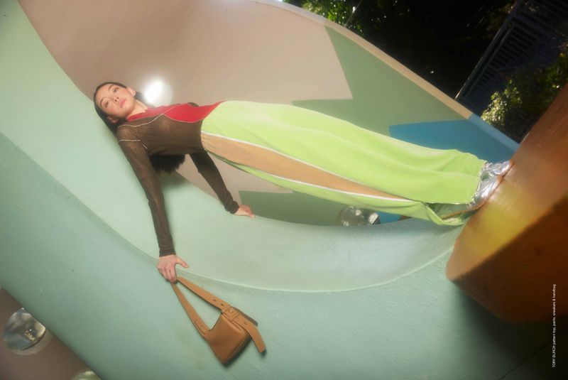 Woman in a fashion pose reclining in a playground structure, holding a purse, wearing a long-sleeved top, green pants and metallic shoes.