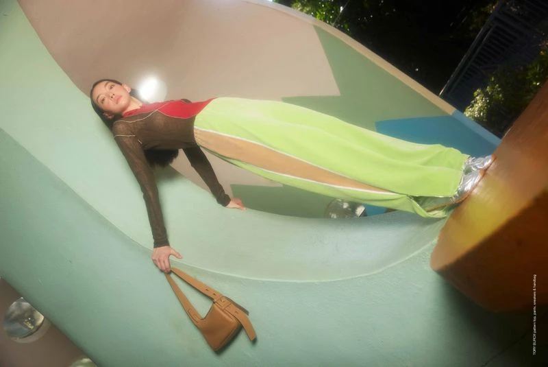 Woman in a fashion pose reclining in a playground structure, holding a purse, wearing a long-sleeved top, green pants and metallic shoes.