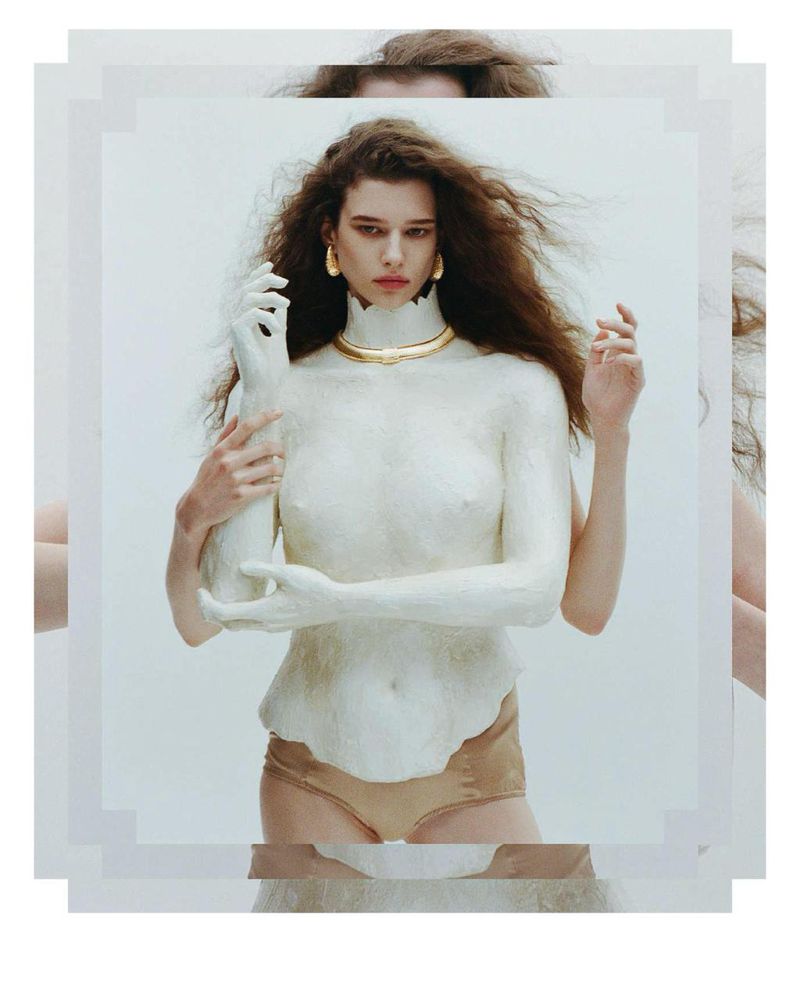 Portrait of a woman with long brown hair and gold jewelry, posing in front of a white background, partially covered in white material.