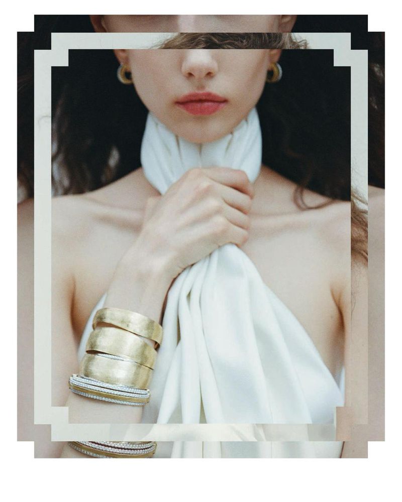 Close-up of a woman with a fair complexion, wearing gold bracelets, a white top, and hoop earrings, set against a dark background.