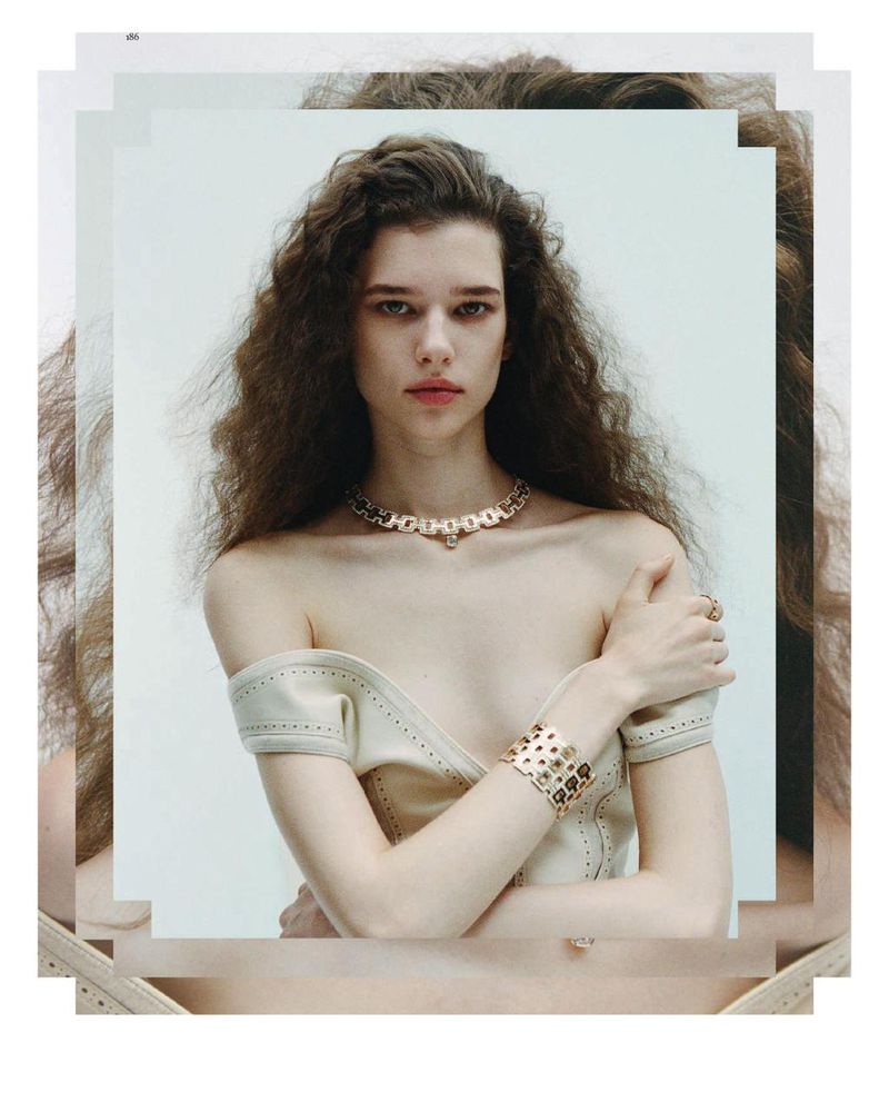 Young woman with long, wavy hair poses wearing a beige dress and gold jewelry against a clean white background in a portrait shot.