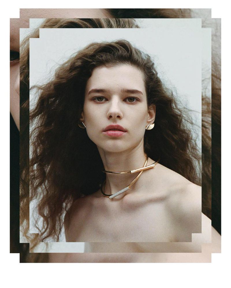 Portrait of a woman with long dark wavy hair wearing a gold choker necklace and earrings against a white background