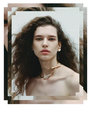 Portrait of a woman with long dark wavy hair wearing a gold choker necklace and earrings against a white background