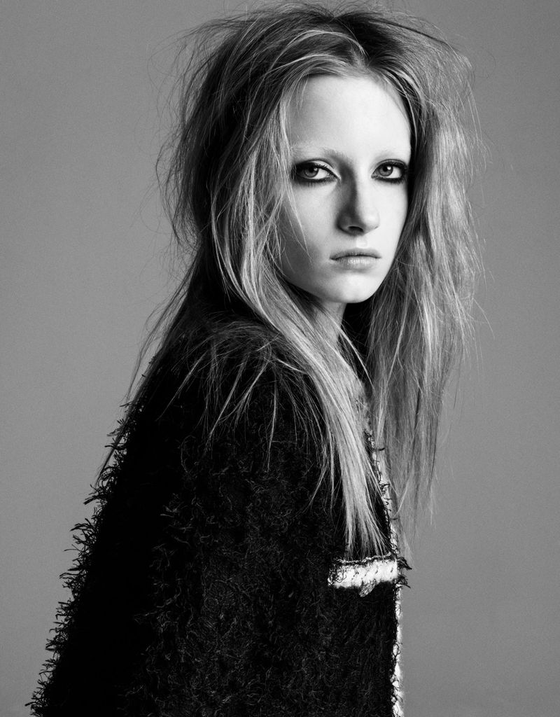 Black and white portrait of a young woman with fair skin, light blonde hair and a dark textured jacket, posing in front of a neutral background.