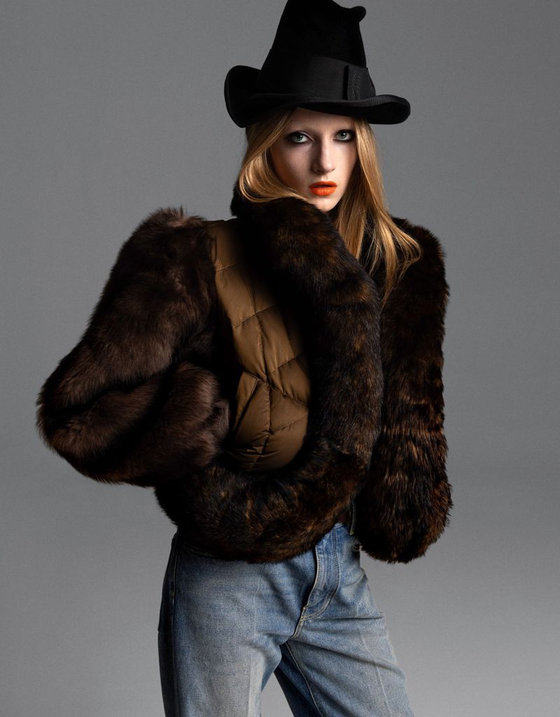 Fashionable woman poses in a fur-trimmed jacket, blue jeans, and a black hat against a grey backdrop in a fashion editorial studio shot.