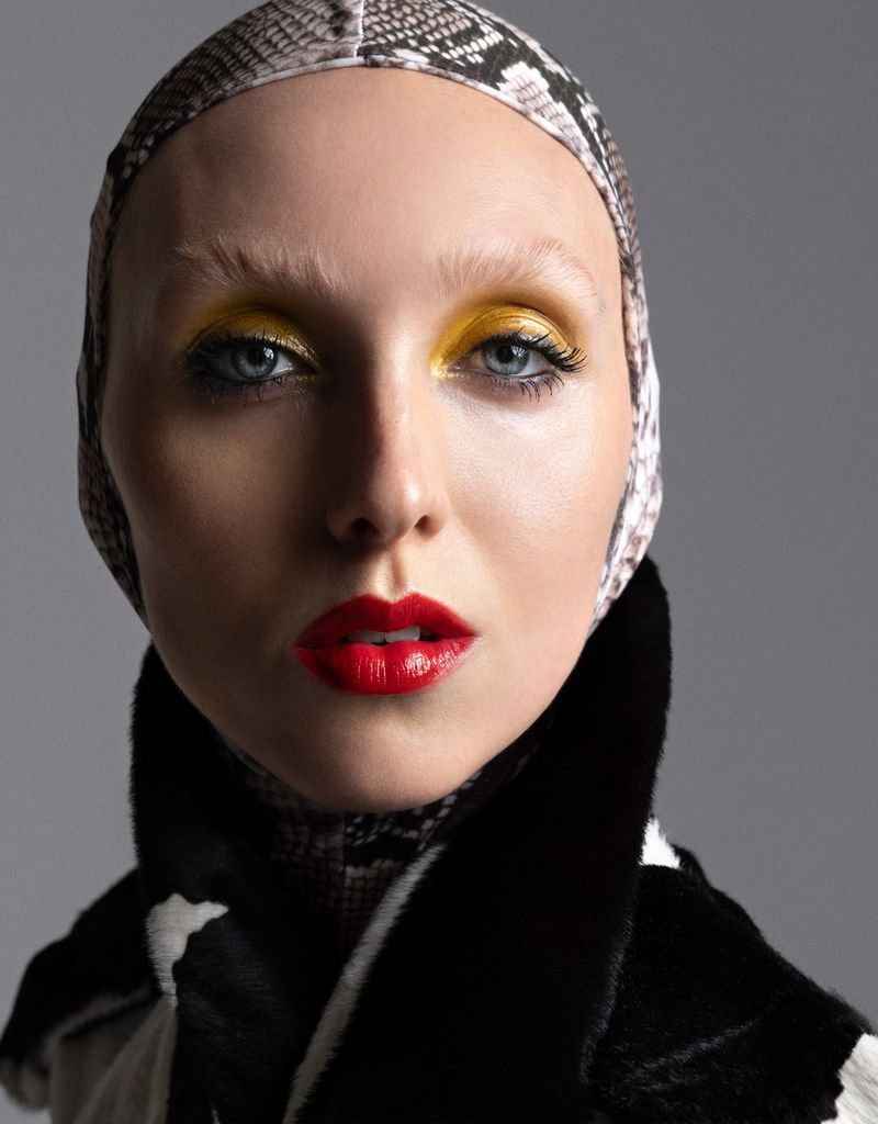 Close-up portrait of a woman with bright yellow eyeshadow and red lipstick, wearing a patterned head covering and a fur-trimmed coat.